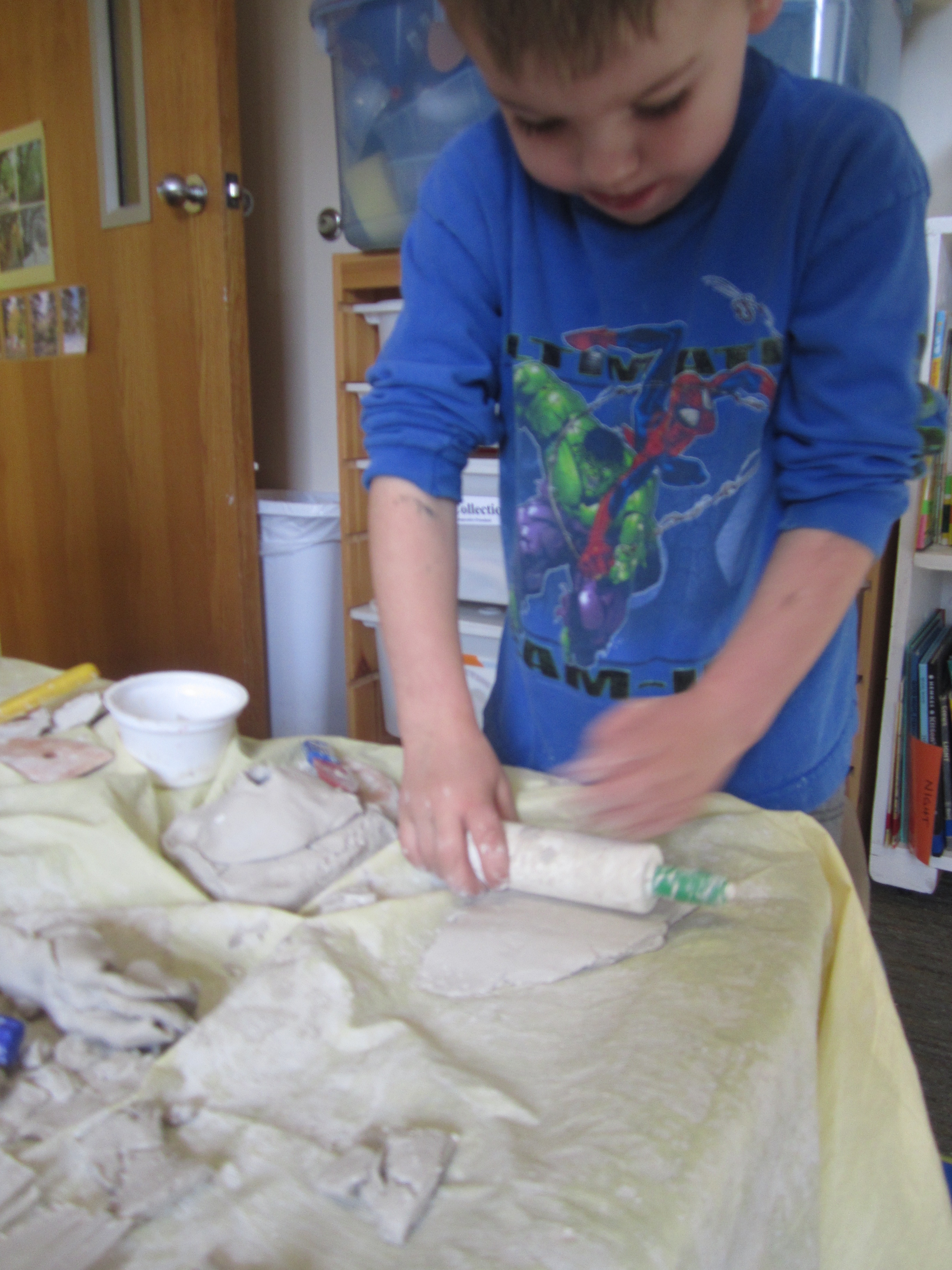 Child uses a small rolling pin to flatten clay.